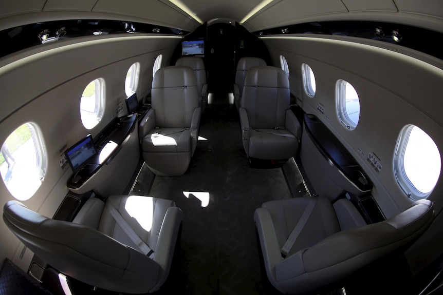 An internal view of a small private jet, showing six leather seats, wooden sideboards and screens