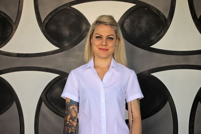 A woman with dyed blonde hair and tattoos stands in front of a patterned wall 