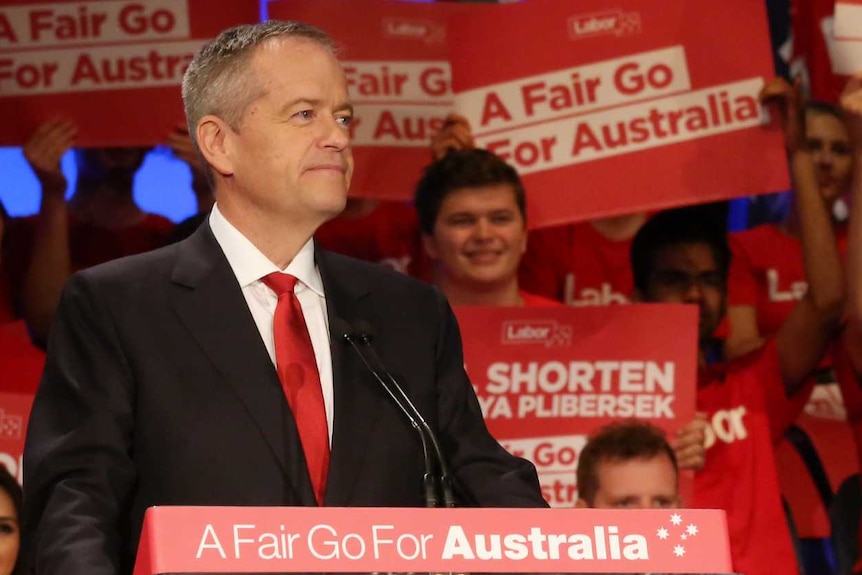 Bill Shorten stands at a lectern with "A Fair Go For Australians" written on it, smiling proudly in front of volunteers