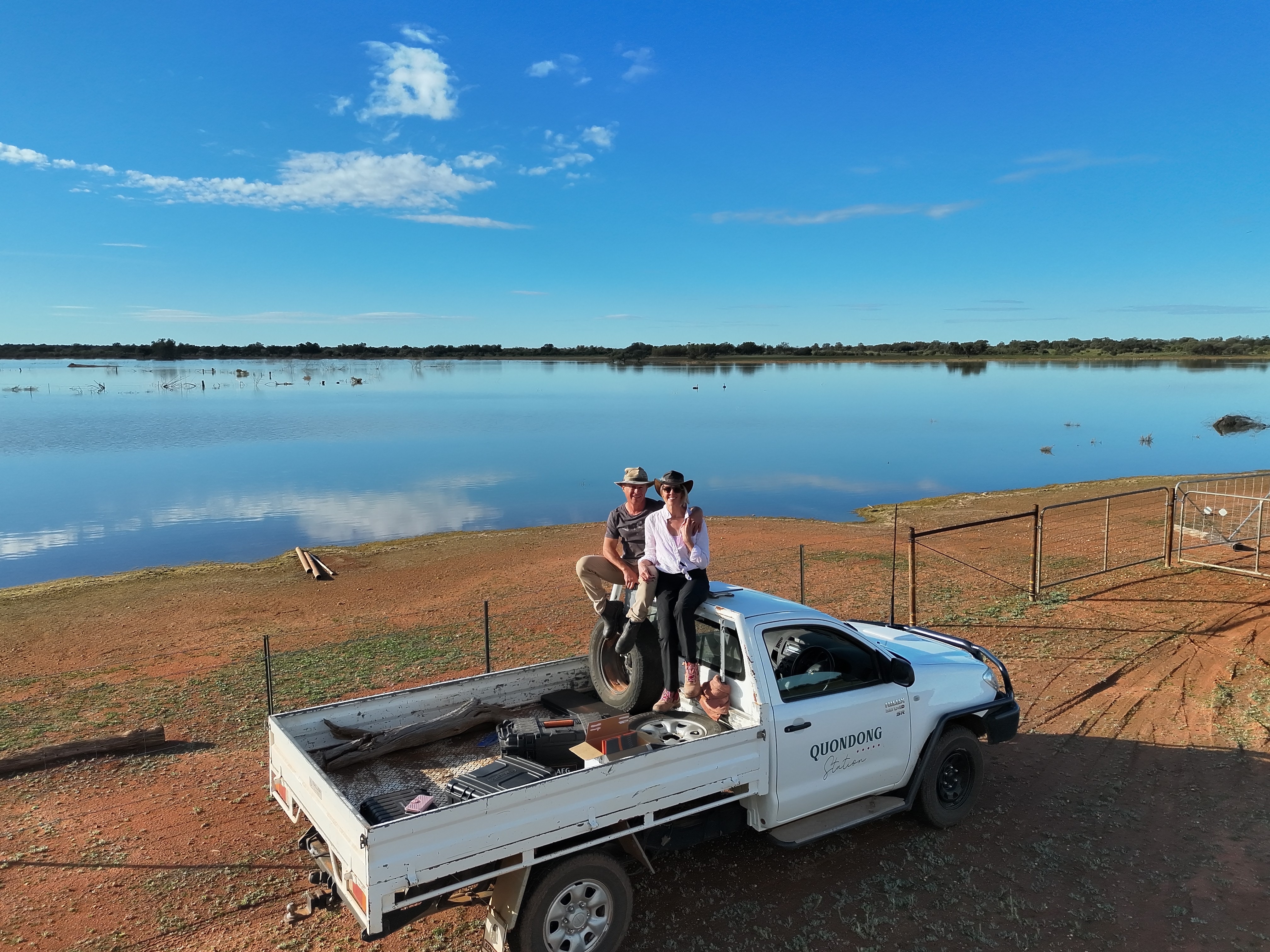 A man and woman sitting ontop of a ute which is parked next to a large body of water on a farm