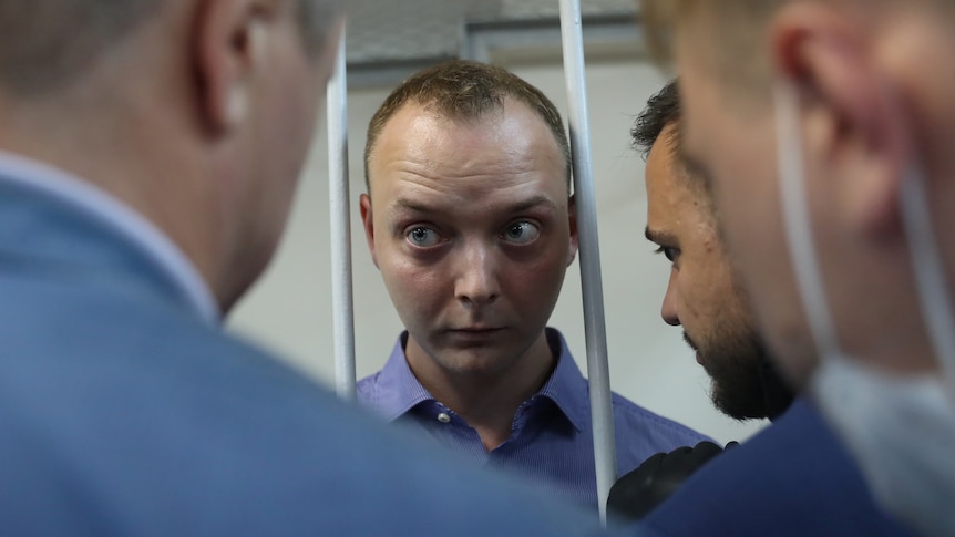 A closeup photo of a man's face between two bars speaking to several other man.