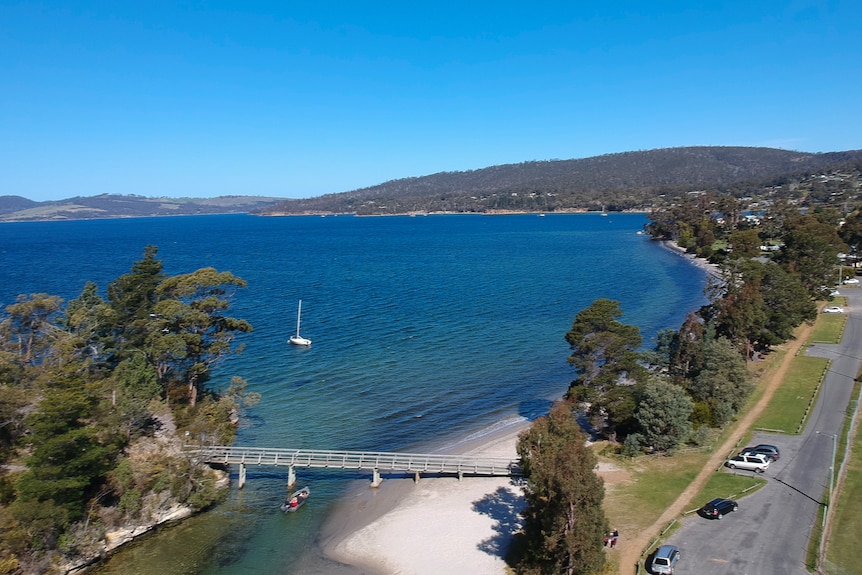 A river reaches the beach. A small watercraft is going under a bridge while another boat is anchored offshore.