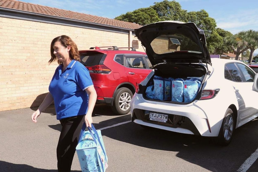 A woman in a polo shirt carries a padded thermal grocery bag from the back of a car boot containing many similar bags.