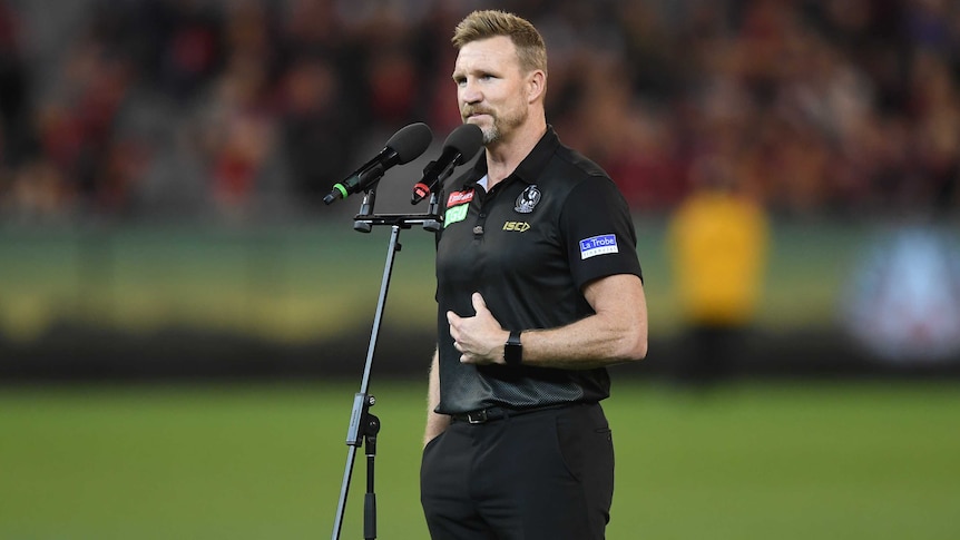 Nathan Buckley stands behind a microphone wearing a black shirt and trousers, with his hand bent across his stomach