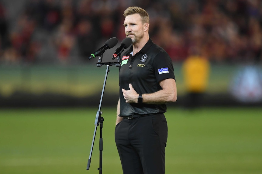 Nathan Buckley stands behind a microphone wearing a black shirt and trousers, with his hand bent across his stomach