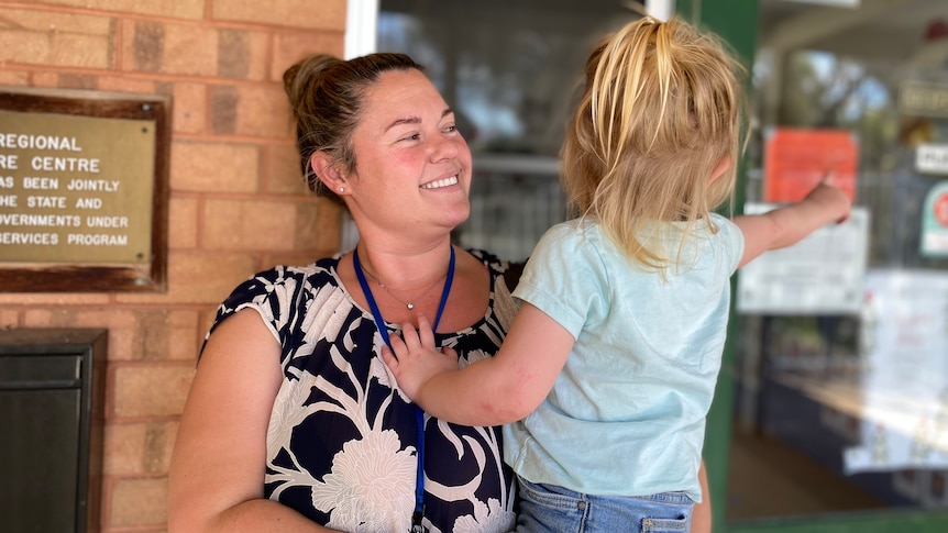 A woman with light brown hair is smiling as she holds a young child wearing a blue shirt with blonde hair.