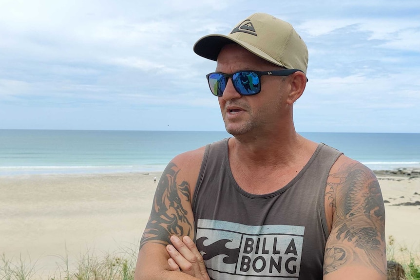 A man wearing sunglasses, a cap and a singlet stands with his arms folded with a beach in the background.