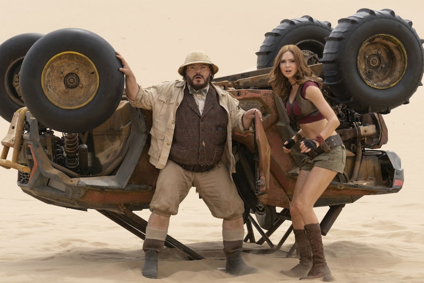 A man dressed as a colonial professor and a woman look startled in front of an upside down desert vehicle on sand
