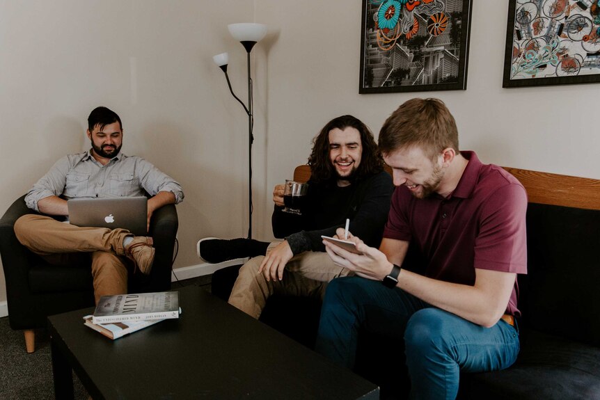 Three men sit in a living room, smiling, drinking coffee and working on a laptop.