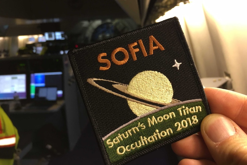 A hand holds a cloth badge that reads "SOFIA Saturn's moon Titan Occultation 2018" with a picture of Saturn and a star on it.