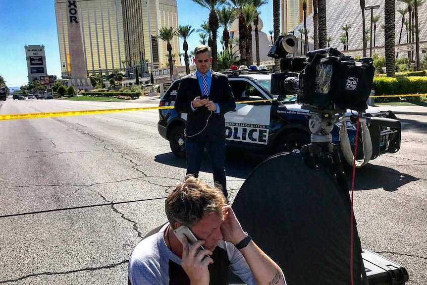 Duffy standing on road in front of police car and tape looking at phone with camera in foreground and cameraman on phone.