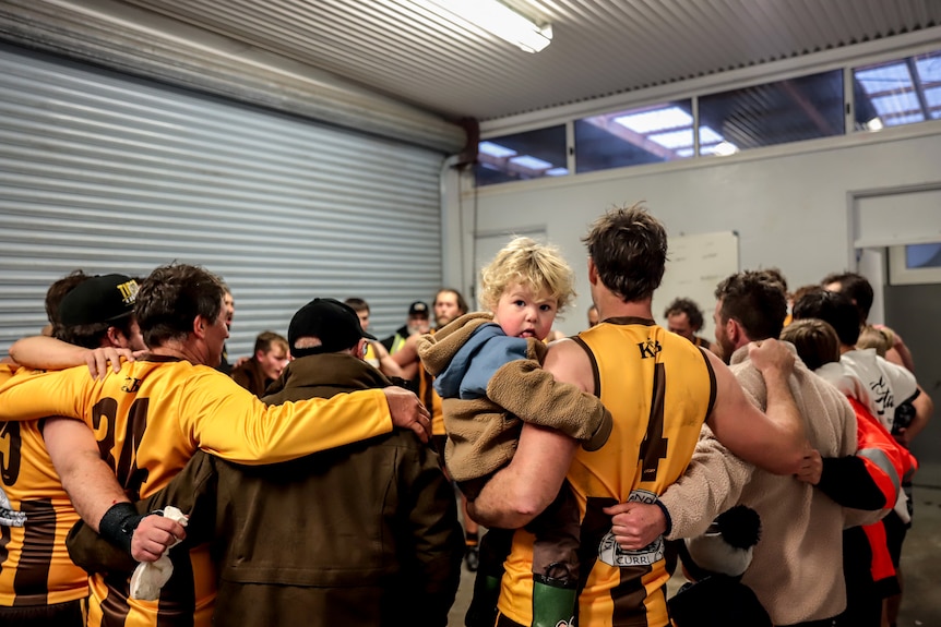 Male football players wearing gold and brown jerseys in fluorescent changerooms with a child on a shoulder