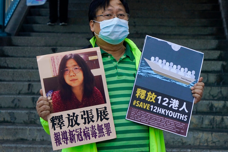 A man in a horizontally-striped polo shirt stands in front of a stairwell, holding two placards related to Chinese detainees.