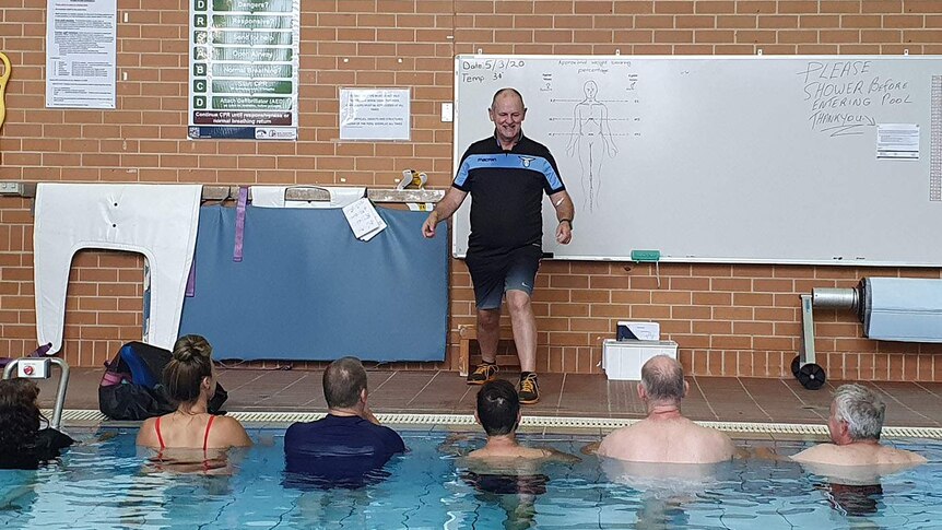 Male instructor standing next to pool looks down at six patients in an indoor swimming pool.