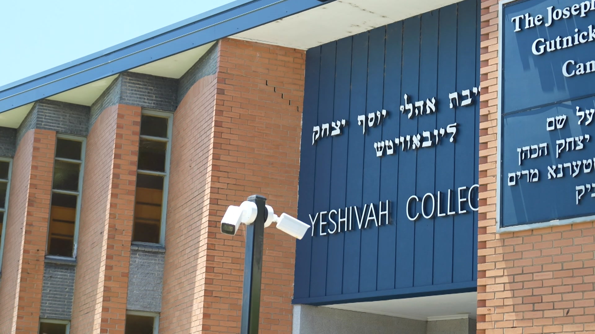 Duas câmeras de segurança brancas estão fixadas em um poste em frente a um prédio de tijolos com uma placa azul que diz "Faculdade Yeshiva".