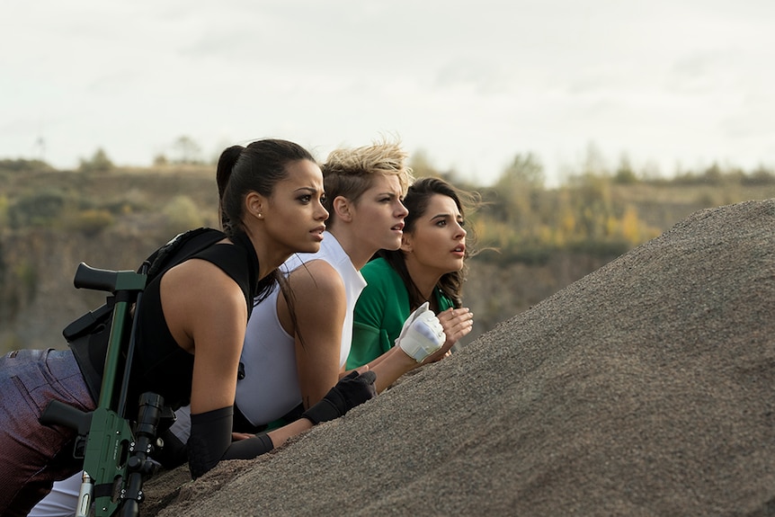 Ella Balinska, Kristen Stewart, and Naomi Scott resting on rock in desert terrain, all three focusing on something in distance.