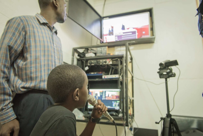 Child speaks on microphone in front of video camera and TV monitor
