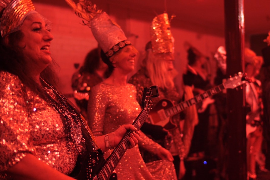 Women with guitars dressed in shiny costumes and headdresses on stage under bright coloured lights, smiling.
