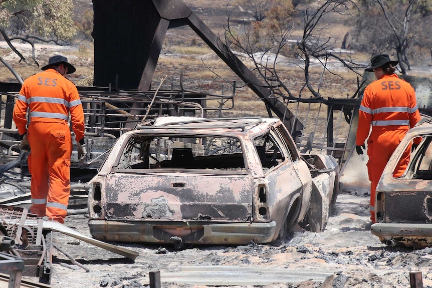 Two men in orange suits inspect a series of burnt out cars.