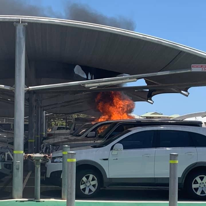 Flames can be seen coming from a car parked in a car park under a shade clothe. There are several cars surrounding it.