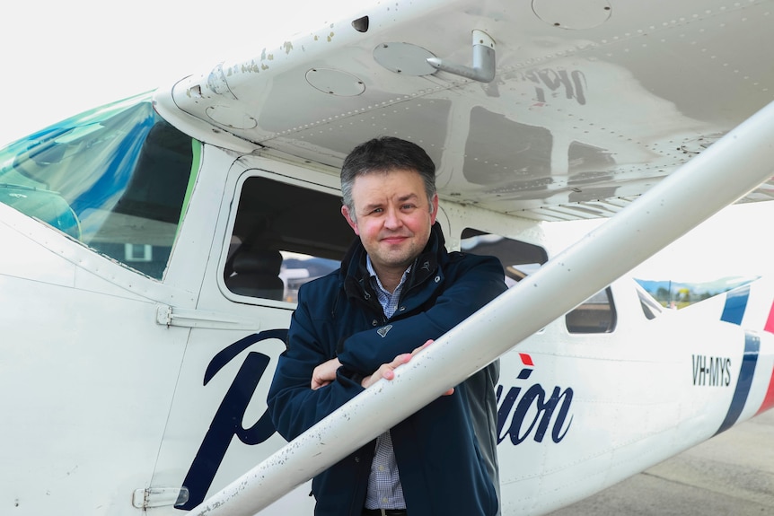 A middle-aged man with a short haircut rests his arms on the wing of a light plane