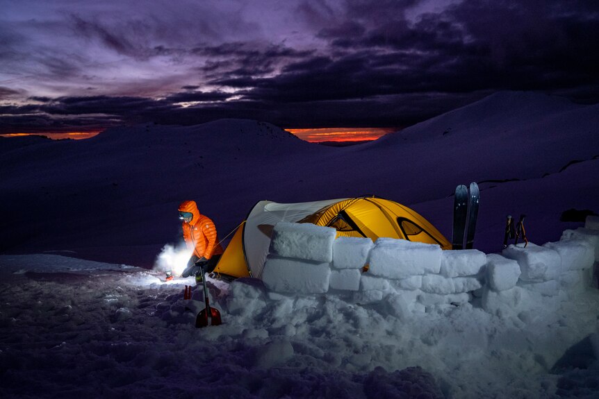 A man uses iceblocks to block the wind from is tent while camping atop a mountain.