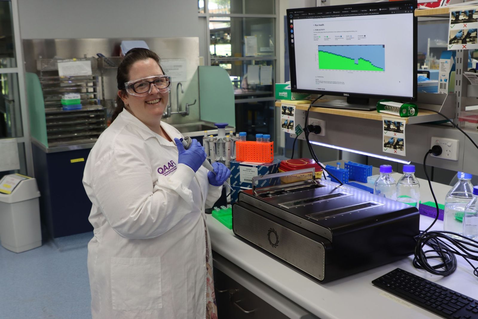 A woman in a white lab coat with safety glasses on holding science equipment in a laboratory.