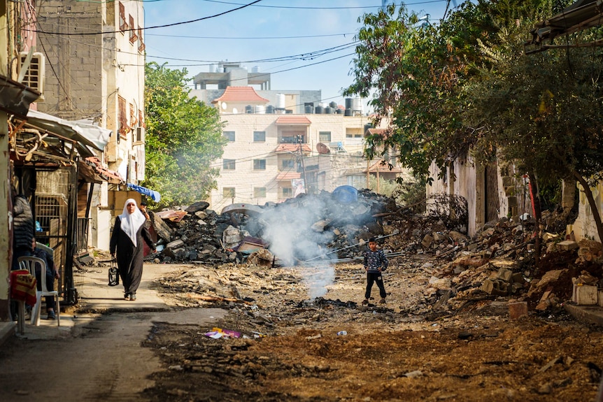 A woman dressed in a hijab walks down a street with smoke rising at the centre of the road.