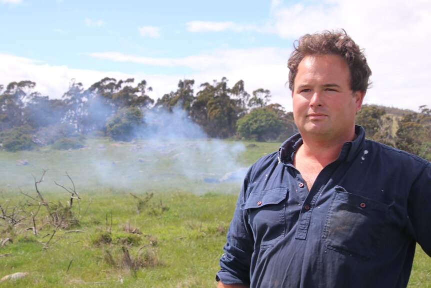 A man in a blue shirt with brown hair stands in front of a trail of smoke, a strand of trees is in the background.  