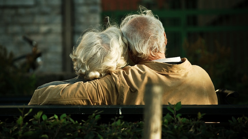 An elderly couple sits on a park bench.