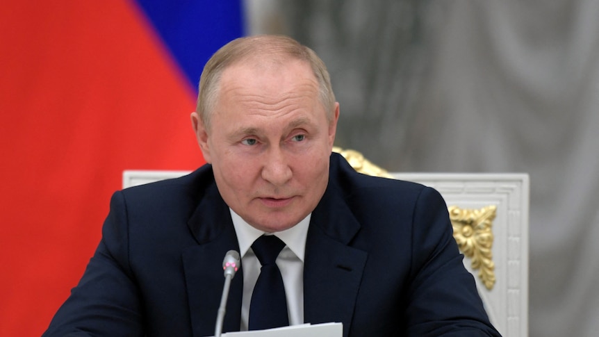 Vladimir Putin, wearing a dark suit, sits behind a microphone holding pieces of paper.