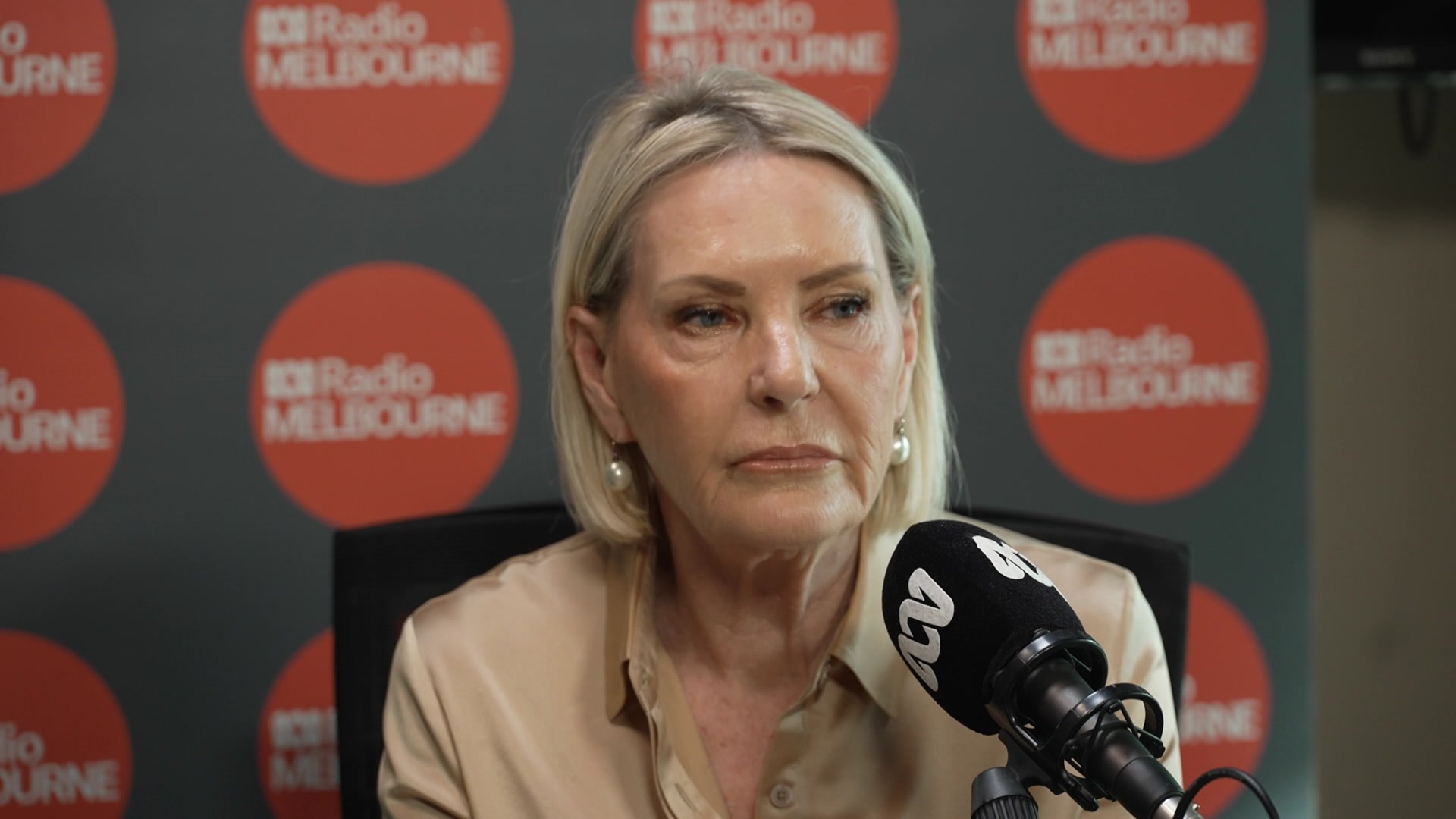 A woman with chin length blonde hair, white pearl earrings in a cream top sits in front of a backdrop with ABC logos.