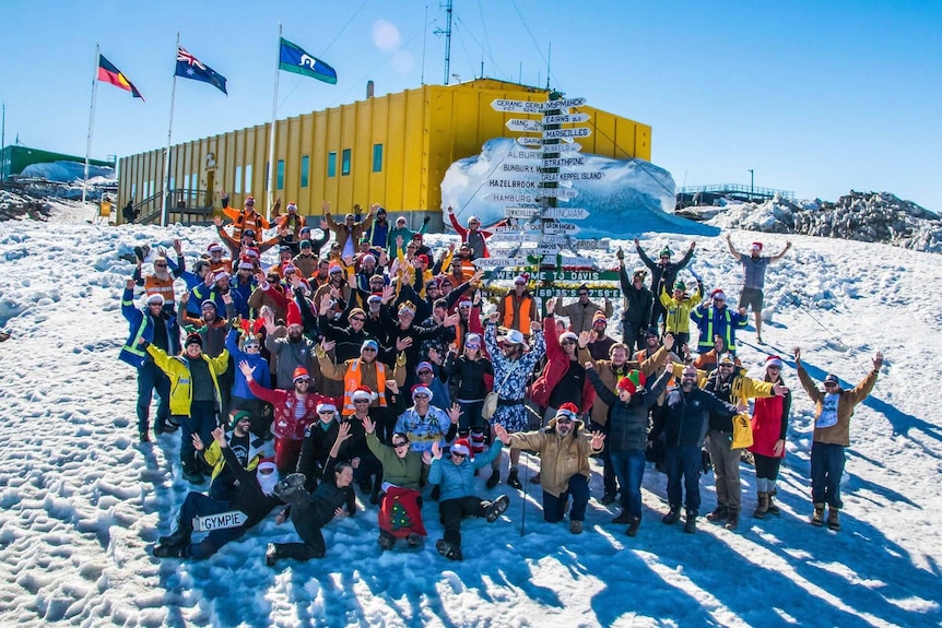 A group of people sit in the snow and wear Santa hats in front of a sign pointing to different cities and a yellow building.