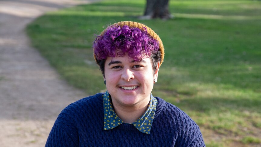 A young person with purple curly hair and a mustard beanie, stands in front of a grassy field, smiling.