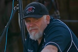 A man with cap and white beard at a racing stables leans on a fence.