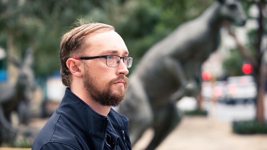 Man with glasses stands in a park with a kangaroo statue behind him.