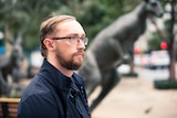 Man with glasses stands in a park with a kangaroo statue behind him.