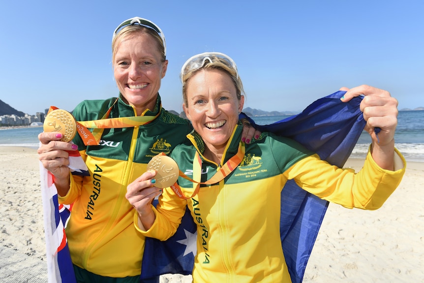 A para-triathlete and her sighted guide stand on a beach in Brazil with their gold medals from the Paralympics.