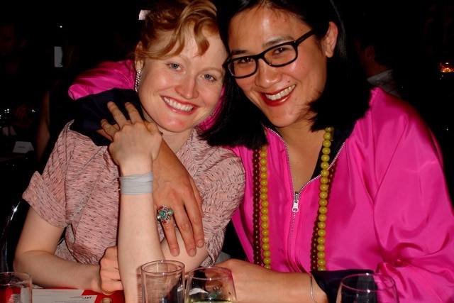 Two women smile big their arms around each other sitting at a table