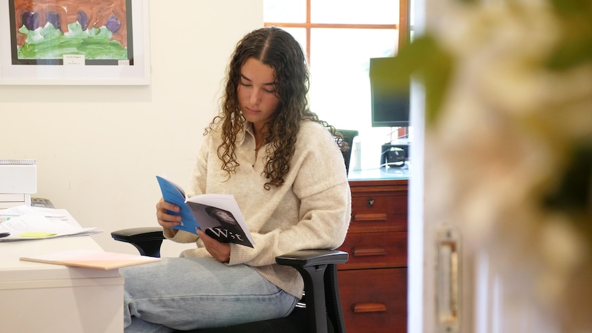 Sheridan sits at her desk wearing a white jumper and jeans and reads a book.