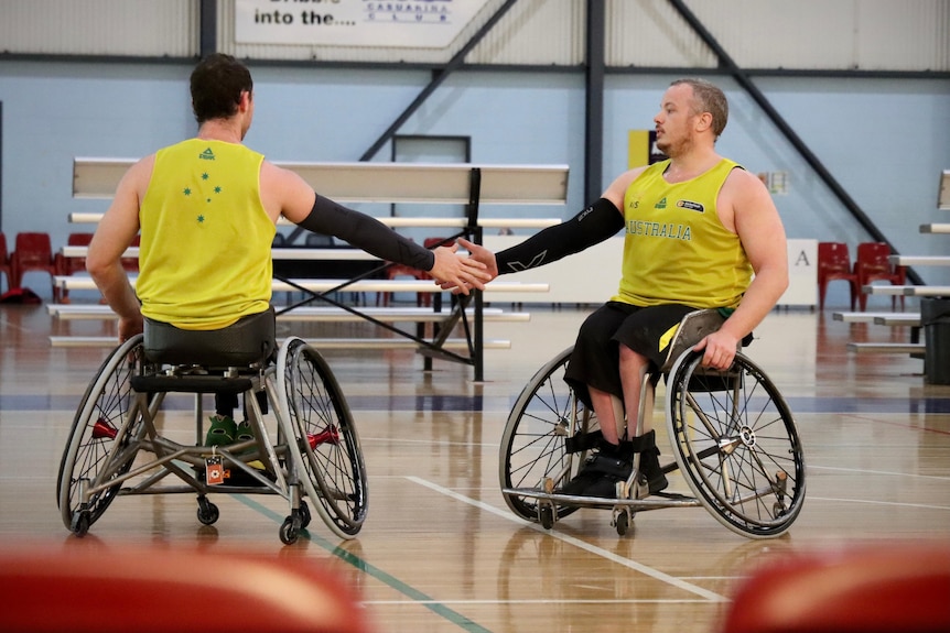 Two young men in wheelchairs wearing green and gold shake hands.