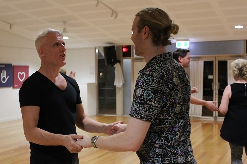 Dance teacher Kevin Flowers teaching a fellow male dancer in a white walled hall.