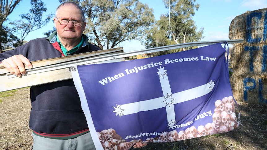 An older man with white hair and glasses holds a Eureka Stockade flag