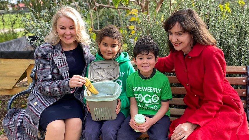 Two women sit on a wooden bench with two children in bright green tshirts, holding a small bin