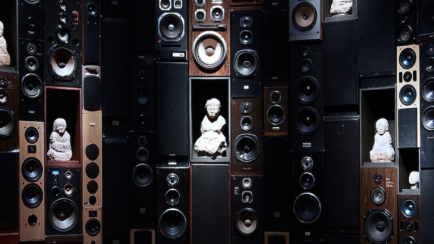 Wall of speakers embedded with small human-shaped stone figures in Buddhist postures.