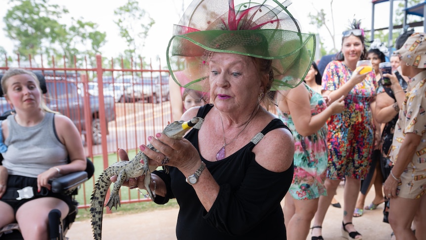 A woman wearing a fascinator holding a juvenile crocodile in an outback pub