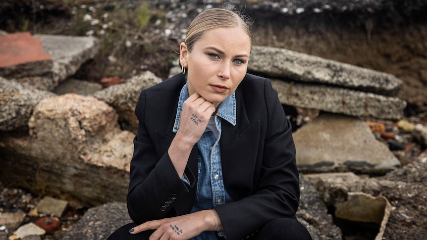 A woman wearing a dark jacket over a blue shirt sits outside and leans her chin on her hand.