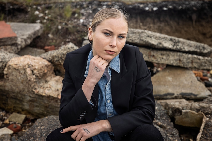 A woman wearing a dark jacket over a blue shirt sits outside and leans her chin on her hand.