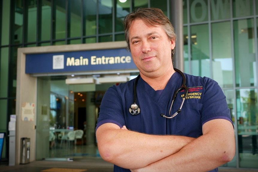 A man wearing hospital scrubs and a stethoscope stands outside the main entrance of a hospital
