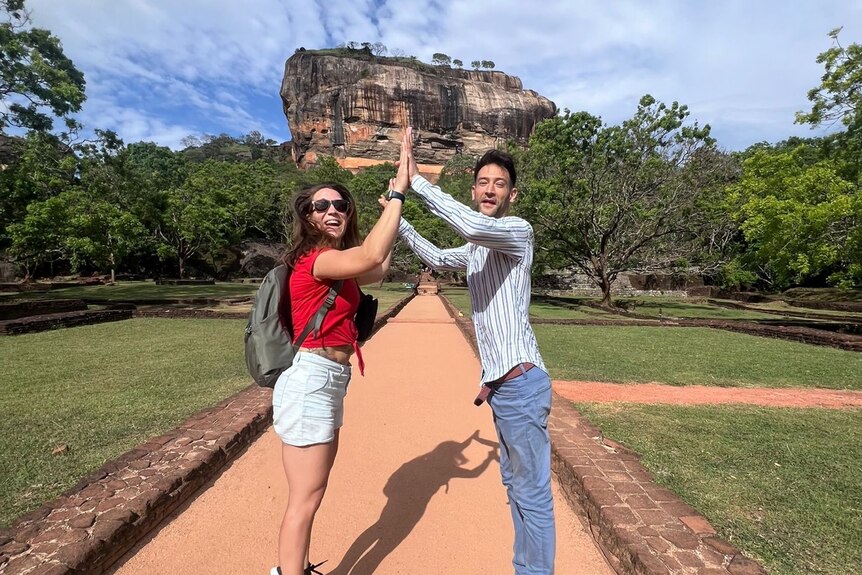 A man and a woman jump and high 5 in front of a rock.